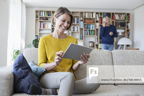 Lächelnde Frau sitzt auf der Couch mit digitalem Tablett,  während ihr Mann im Hintergrund telefoniert.