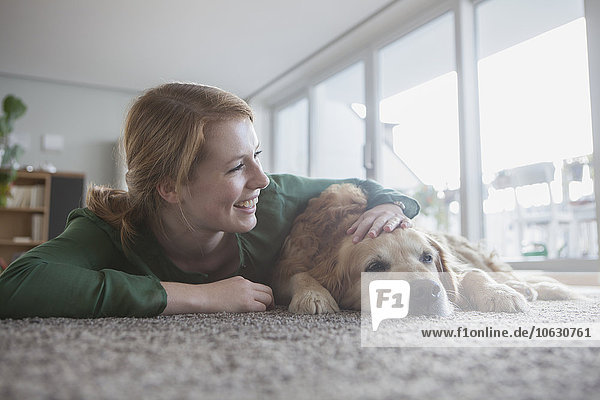Lächelnde junge Frau neben ihrem Hund auf dem Teppich liegend