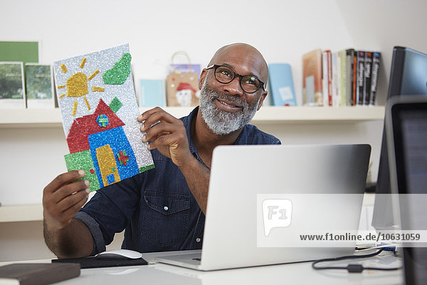 Portrait of smiling man sitting at his desk showing child's drawing