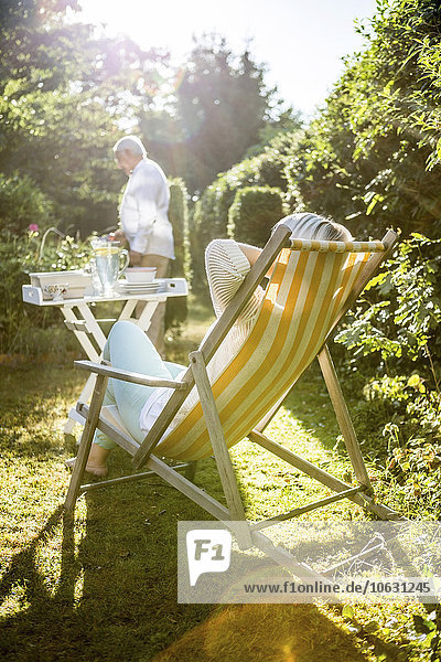 Reife Frau entspannt im Liegestuhl im Garten mit Mann im Hintergrund
