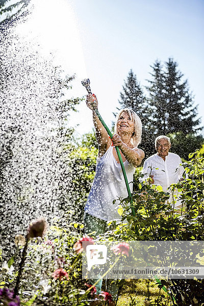 Lächelnde reife Frau beim Blumen gießen im Garten mit Mann im Hintergrund