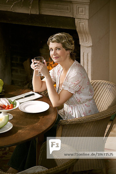 Porträt einer lächelnden reifen Frau  die am gedeckten Tisch sitzt und ein Glas Rotwein hält.