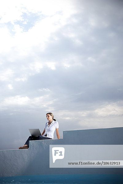 Spanien  Mallorca  Frau mit Laptop auf der Treppe vor bewölktem Himmel sitzend