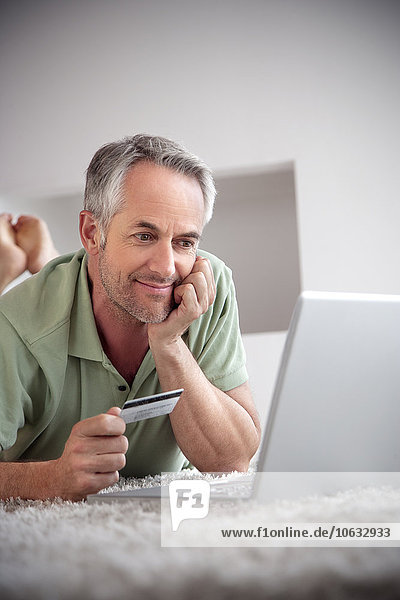 Porträt eines lächelnden Mannes  der mit Laptop auf einem Teppich mit Kreditkarte liegt.