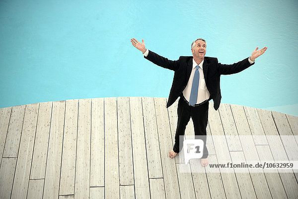 Barefoot businessman wearing black suit standing in front of swimming pool with arms raised
