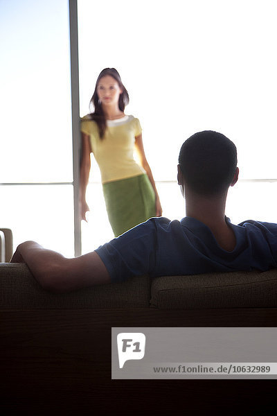 Junger Mann auf der Couch mit Blick auf die Freundin am Fenster
