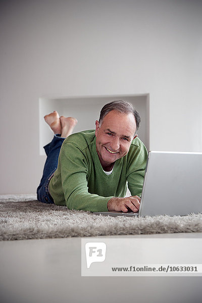 Portrait of smiling man lying with laptop on a carpet at home
