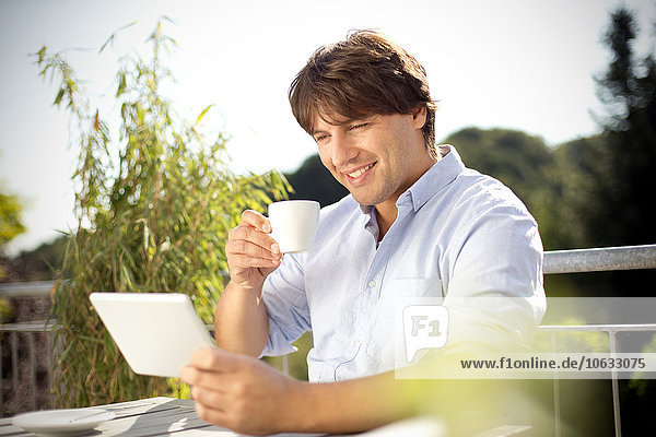 Lächelnder Mann mit digitalem Tablett und Tasse Kaffee auf dem Balkon