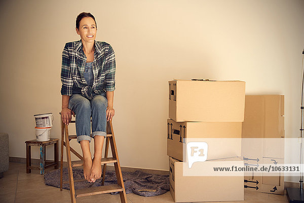 Smiling woman sitting on step ladder beside cardboard boxes