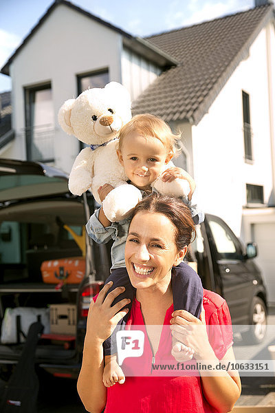 Mother carrying son with teddy bear on shoulders  standing in front of home