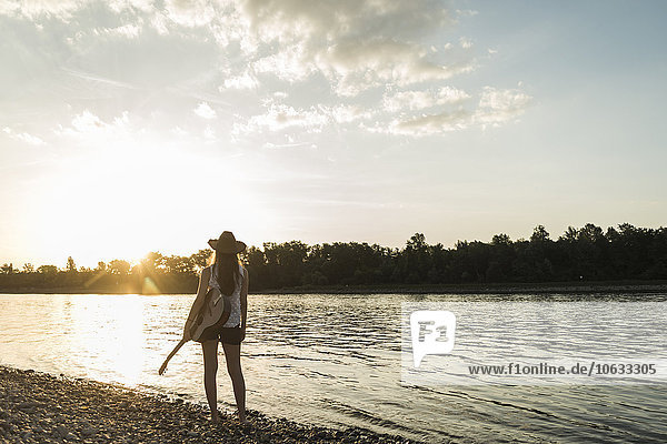 Junge Frau mit Gitarre am Flussufer beim Sonnenuntergang