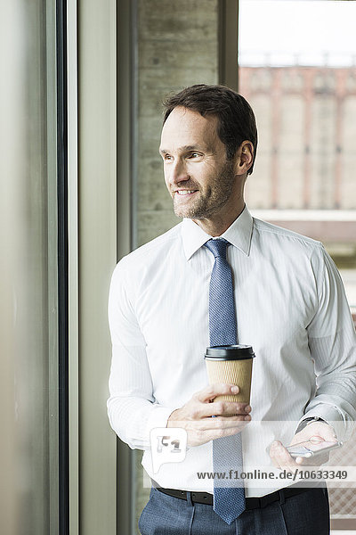 Porträt eines lächelnden Geschäftsmannes mit Kaffee und Smartphone durchs Fenster schauend