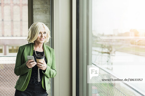 Porträt einer blonden Geschäftsfrau mit Kaffee zum Mitnehmen und Smartphone mit Blick durchs Fenster