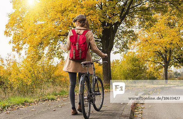 Young woman with backpack pushing her bicycle in autumn landscape