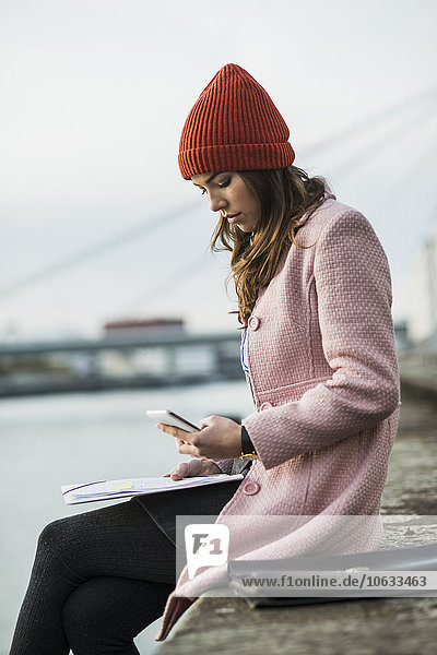 Junge Frau sitzt am Flussufer und schaut auf das Handy
