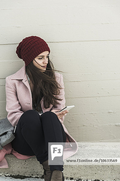 Young woman sitting at concrete wall holding cell phone
