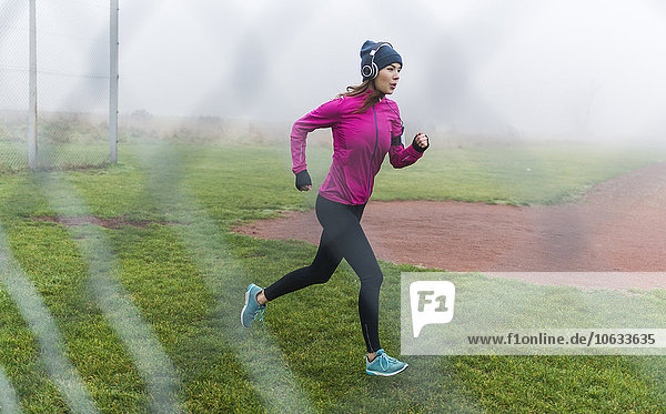 Junge Frau mit Kopfhörer joggen auf einer Wiese hinter Drahtgewebe