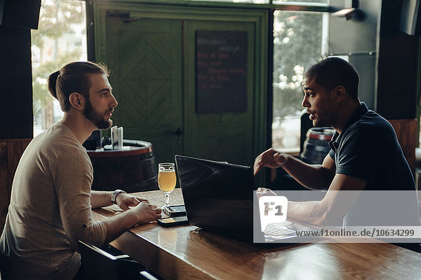 Two men with laptop sitting in a bar talking