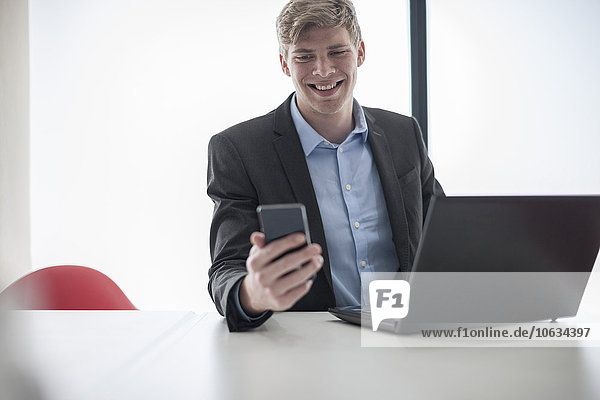Lächelnder Geschäftsmann am Schreibtisch mit Handy und Laptop