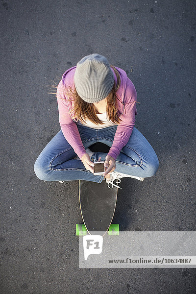 Teenager-Mädchen sitzt auf dem Skateboard und schaut auf das Handy.
