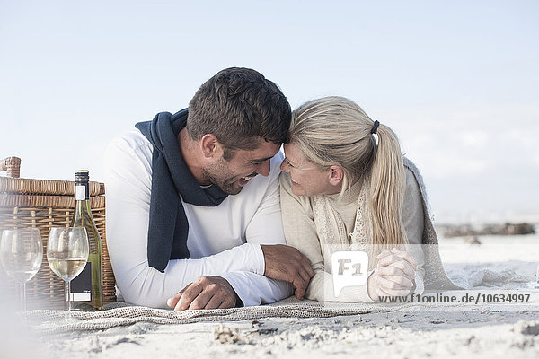Lachendes Paar  das auf einer Decke am Strand liegt und Picknick macht.