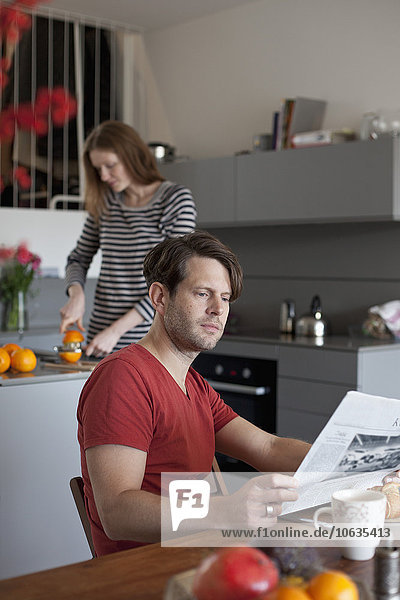 Mann liest Zeitung in der Küche  Frau macht Orangensaft im Hintergrund