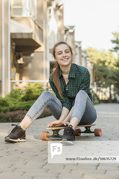 Porträt einer glücklichen jungen Frau  die auf einem Skateboard im Freien sitzt.