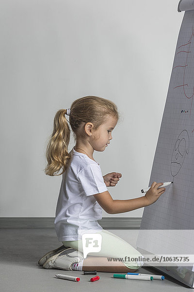 Side view of girl kneeling while drawing on flipchart against white wall