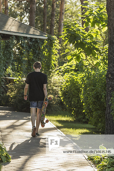 Rear view of man carrying skateboard while walking on footpath at park