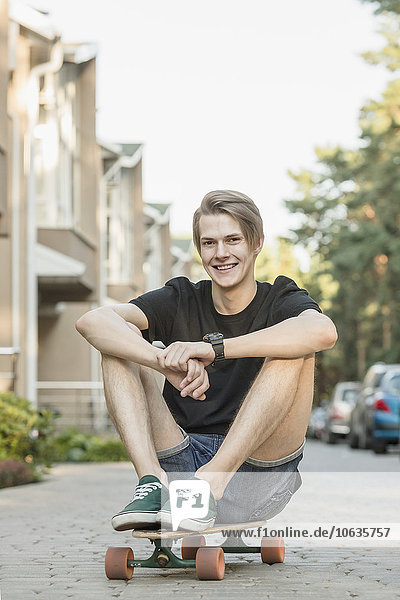 Porträt eines lächelnden jungen Mannes  der auf einem Skateboard im Freien sitzt.