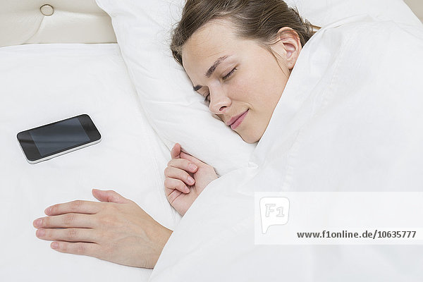 Hochwinkelansicht einer schönen Frau  die mit einem Smartphone auf einem Kissen im Bett schläft.