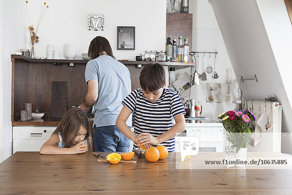 Junge quetscht Orangen  während die Schwester mit dem Vater im Hintergrund in der Küche Saft trinkt.