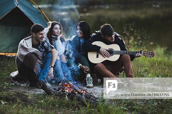 Mann spielt Gitarre  während er mit Freunden am Lagerfeuer sitzt.