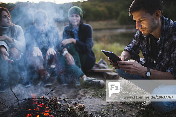 Junger Mann beim Sitzen mit Freunden am Lagerfeuer mit digitalem Tablett