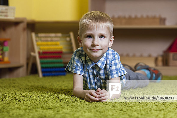 Ganzflächiges Porträt des Jungen  der im Klassenzimmer auf einem Teppich liegt.