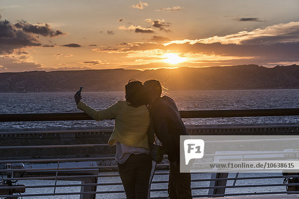 Rear view of couple taking selfie at railing by sea during sunset