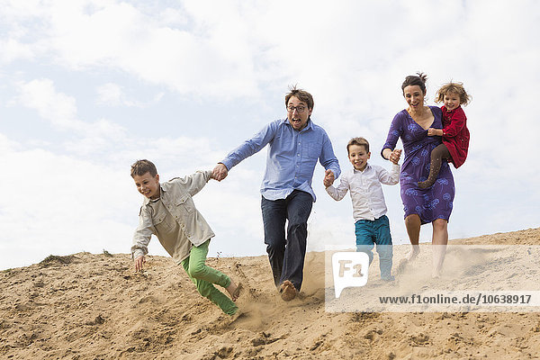 Fröhliche Familie hält sich an den Händen  während sie auf einer Sanddüne gegen den Himmel rennt.