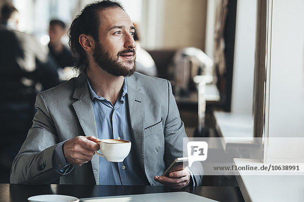 Mid adult businessman holding coffee cup and mobile phone in cafe