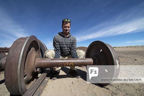 Junger Mann hebt Achse von Eisenbahn wie eine Hantel,  Cementerio de los trenes,  Uyuni,  Departement Potosi,  Altiplano,  Bolivien,  Südamerika