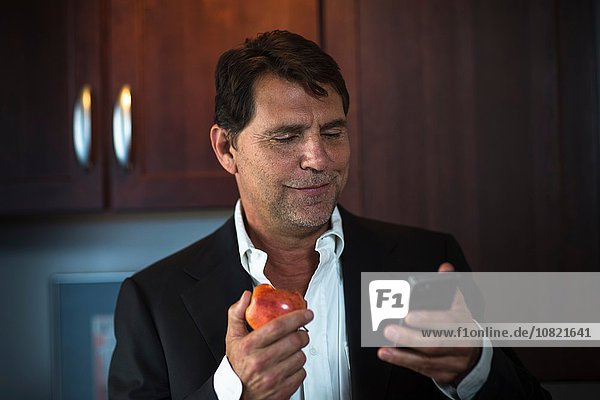 Reifer Mann im Anzug Jacke mit Apfel  Blick auf Smartphone lächelnd