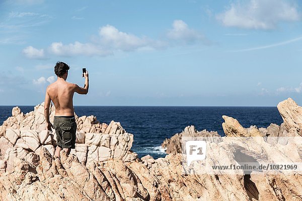 Rückansicht des jungen Mannes auf dem Felsen mit dem Smartphone  um den Horizont über dem Meer zu fotografieren  Costa Paradiso  Sardinien  Italien