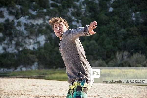 Frontansicht eines jungen Mannes am Strand  der in Wurf-Action steht und lächelnd in die Kamera schaut  Costa Smeralda  Sardinien  Italien