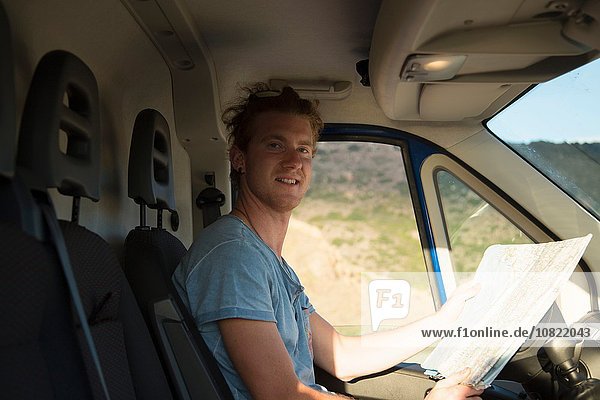 Side view of young man sitting in van holding map looking at camera smiling  Costa Smeralda  Sardinia  Italy