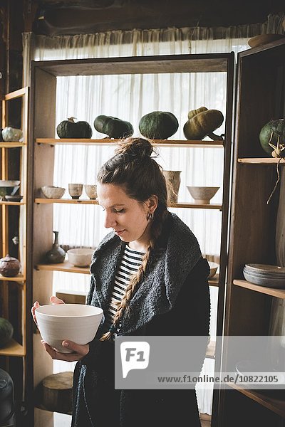 Junge Frau hält Keramikschale vor Regalen mit Tontöpfen und Kürbissen