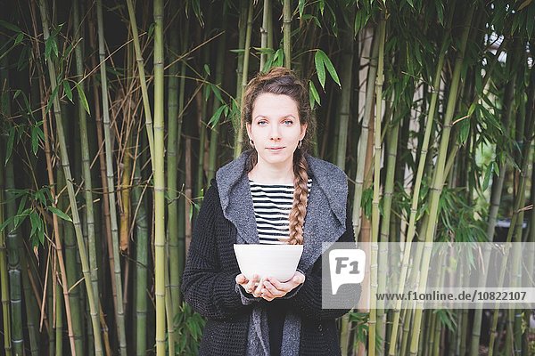 Front view of young woman standing in front of bamboo grove holding ceramic dish looking at camera