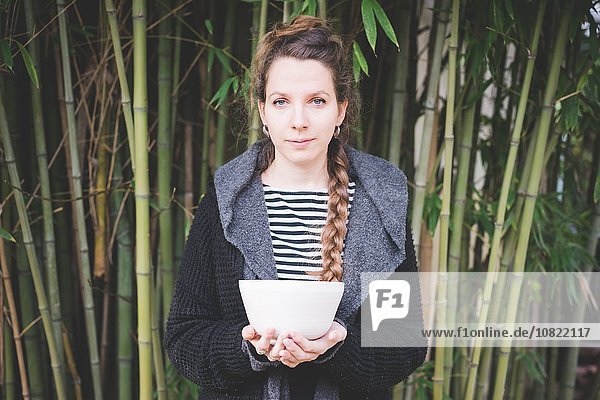 Front view of young woman standing in front of bamboo grove holding ceramic dish looking at camera