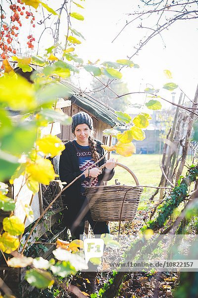 View through leaves of young woman in garden wearing knit hat holding wickerwork basket looking at camera smiling