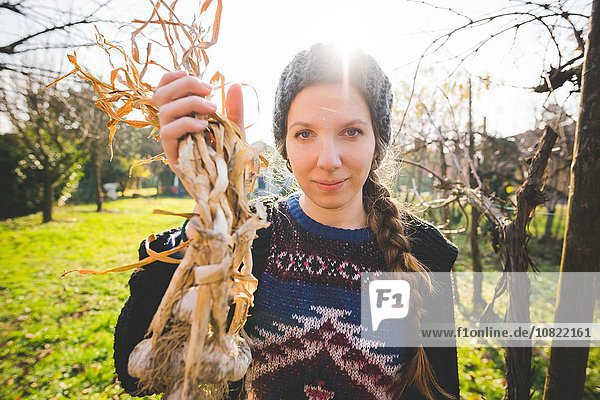Young woman in garden holding freshly picked garlic bulbs looking at camera smiling