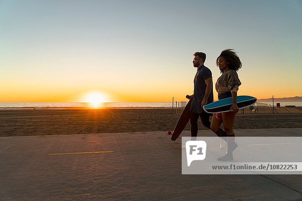 Couple walking along pathway by beach  holding skateboards