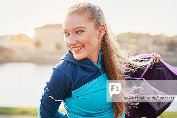 Mid adult female runner putting on tracksuit top at city riverside